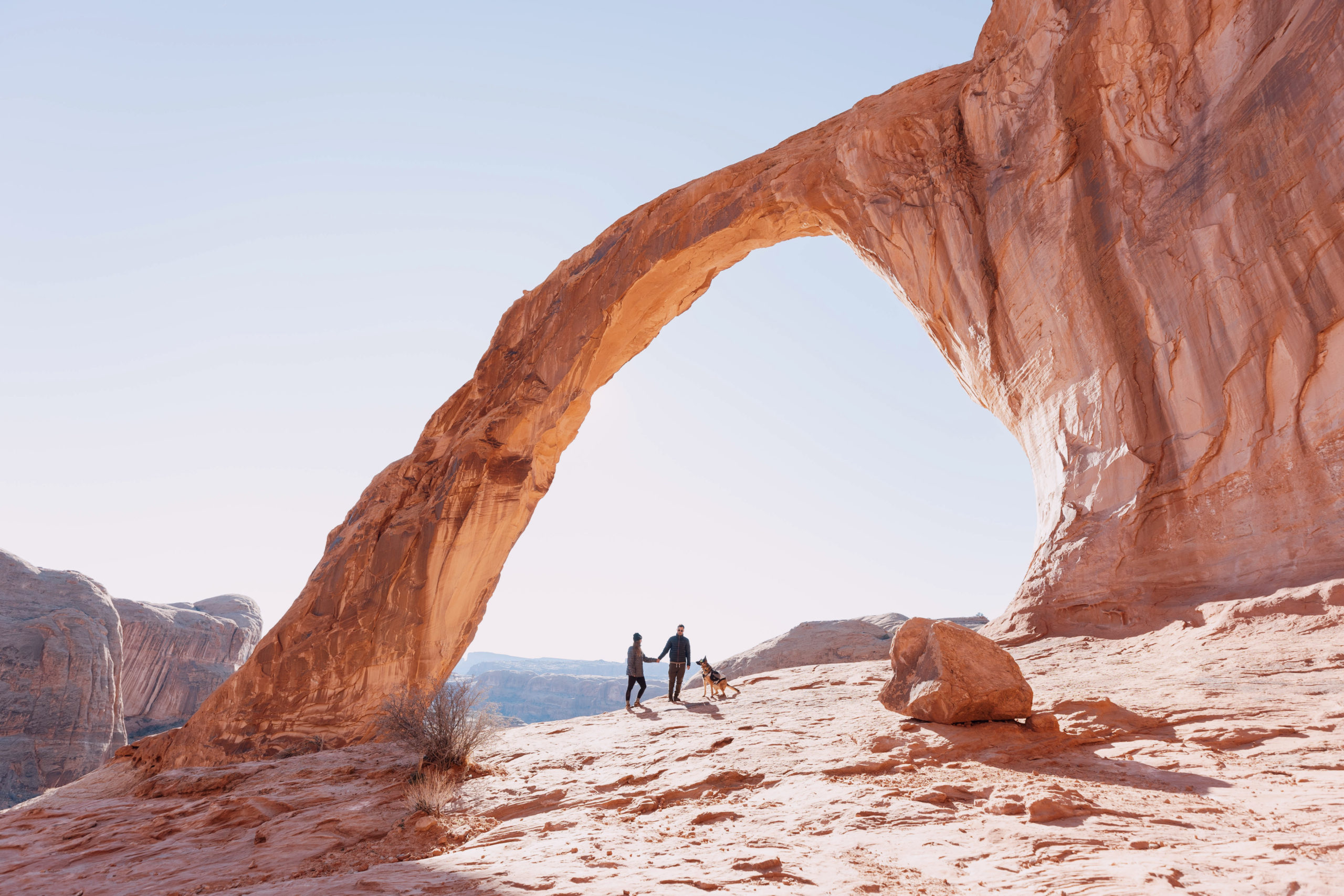 picture of a man, woman, and their dog hiking at corona arch in moab, utah. in the photo, the sun is shining bright behind the arch and the couple is holding hands walking through the center of the arch. the arch is a warm color of sandstone and there are broken pieces of rock in the desert landscape.
